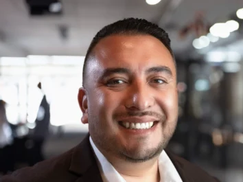 Smiling businessman in a dark suit and white shirt posing in a modern office setting.