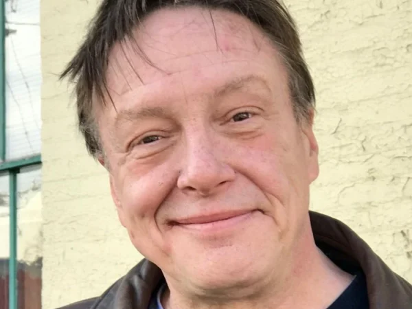 Larry Dunn smiling portrait outdoors wearing brown leather jacket in front of pale brick wall