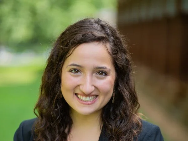 Sophie Mariam professional headshot, smiling with curly brown hair, navy blazer, blurred garden background