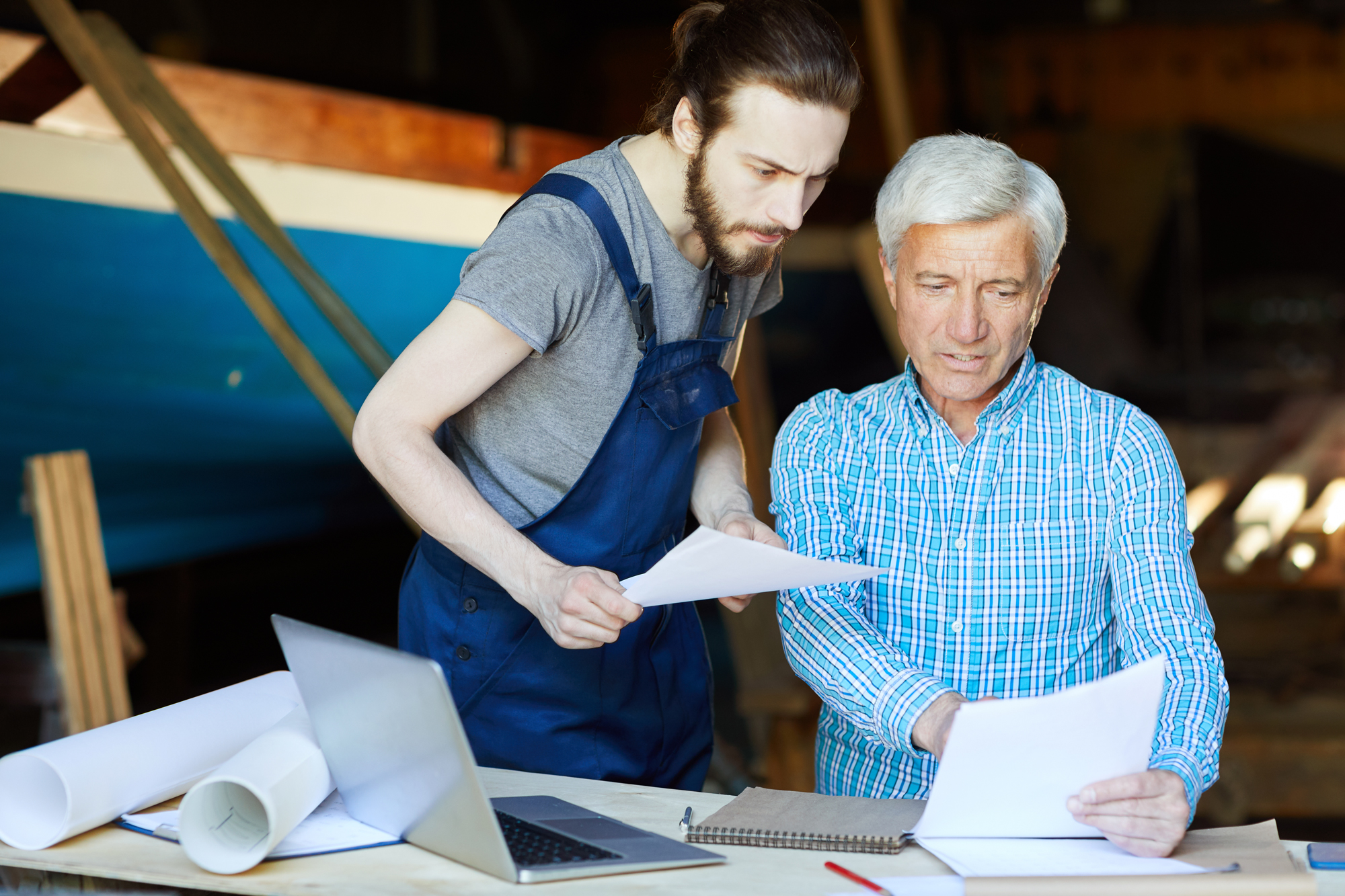 Younger construction worker and older owner reviewing blueprints at workshop table with laptop