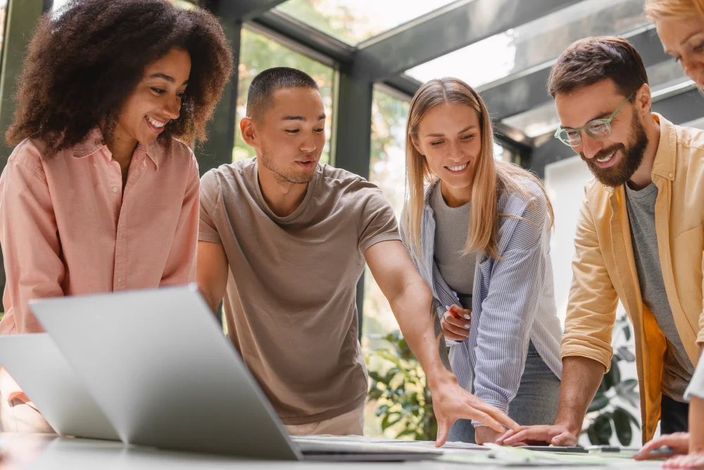 Diverse coworkers collaborating around laptop, pointing at screen and smiling
