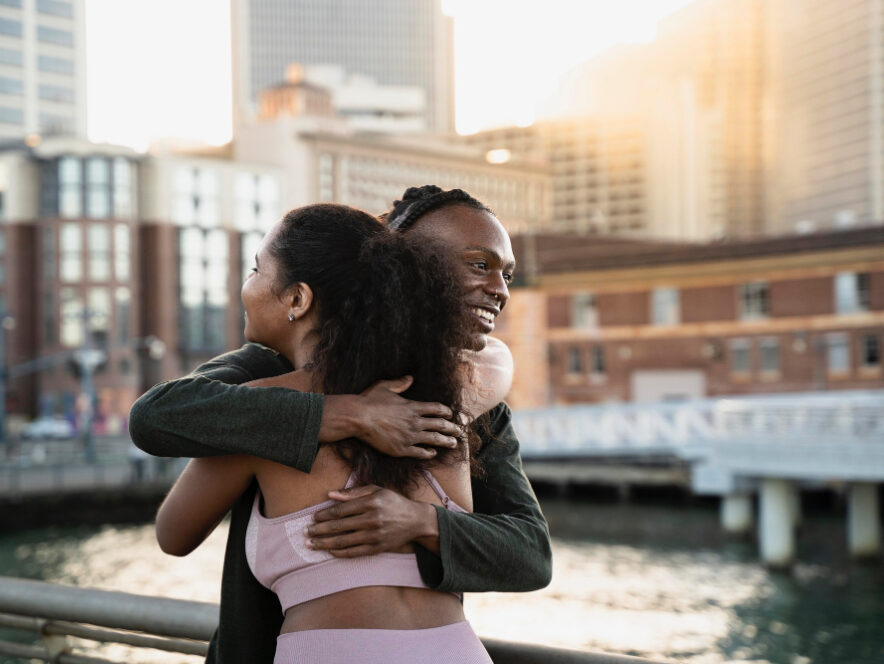 Two friends hugging on waterfront boardwalk with city skyline background