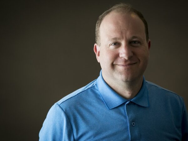 Jared Polis smiling headshot wearing blue polo against dark background