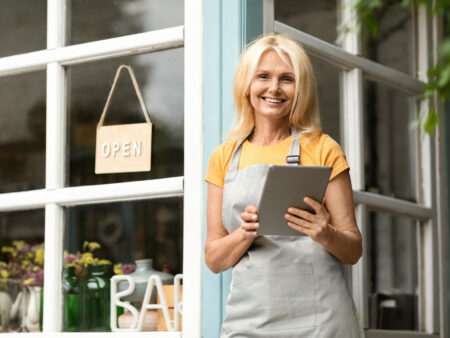 Female shop owner in apron holding tablet at storefront window with open sign
