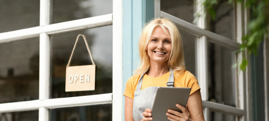 Female shop owner in apron holding tablet at storefront window with open sign
